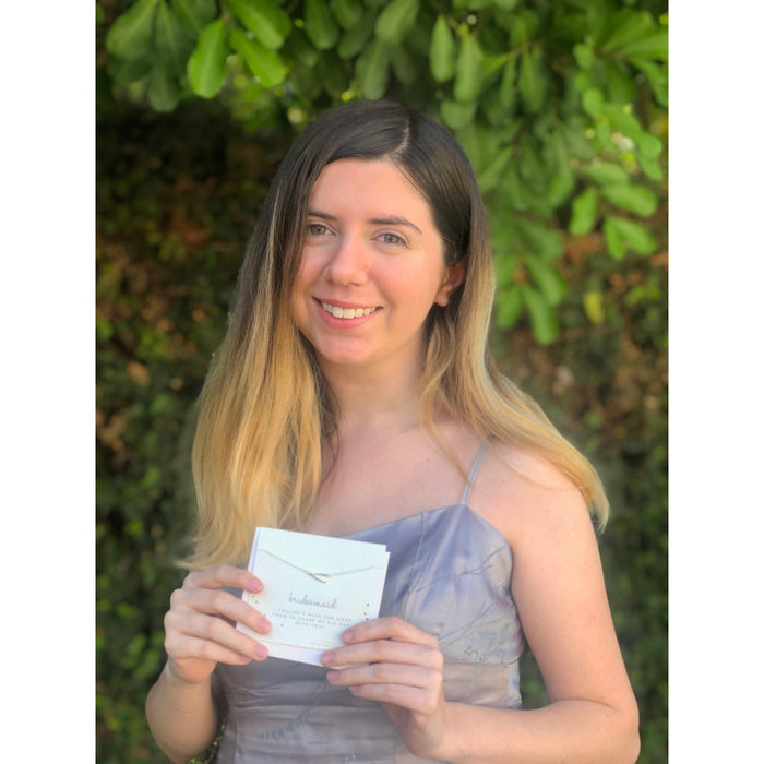 A woman in a dress smiles and holds a card that says 'bridesmaid' with a necklace attached.