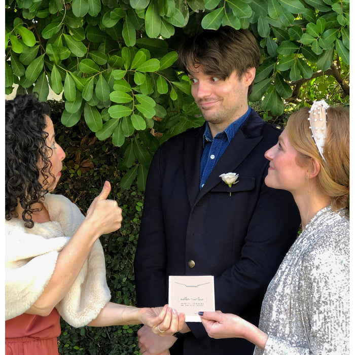 A woman gives a thumbs up while the bride and groom hold a 'mother-in-law' gift card near a leafy green backdrop.
