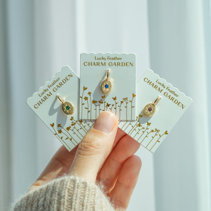 A hand holds three 'Lucky Feather Charm Garden' pendants, each with a different colored gemstone.
