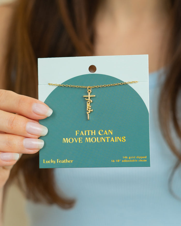 A person holds a teal card displaying a gold 'Faith' necklace and the words 'Faith Can Move Mountains'.