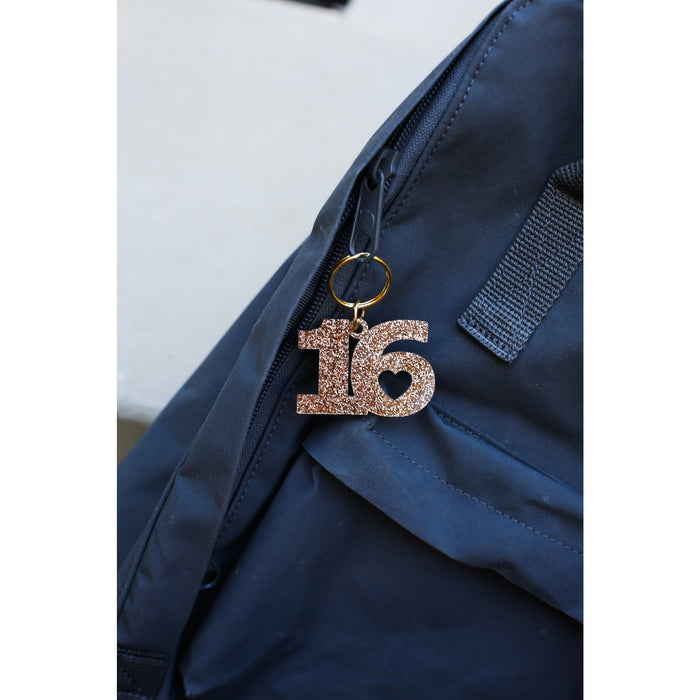 A sparkly, rose gold number 16 keychain with a heart detail hangs from a blue backpack.