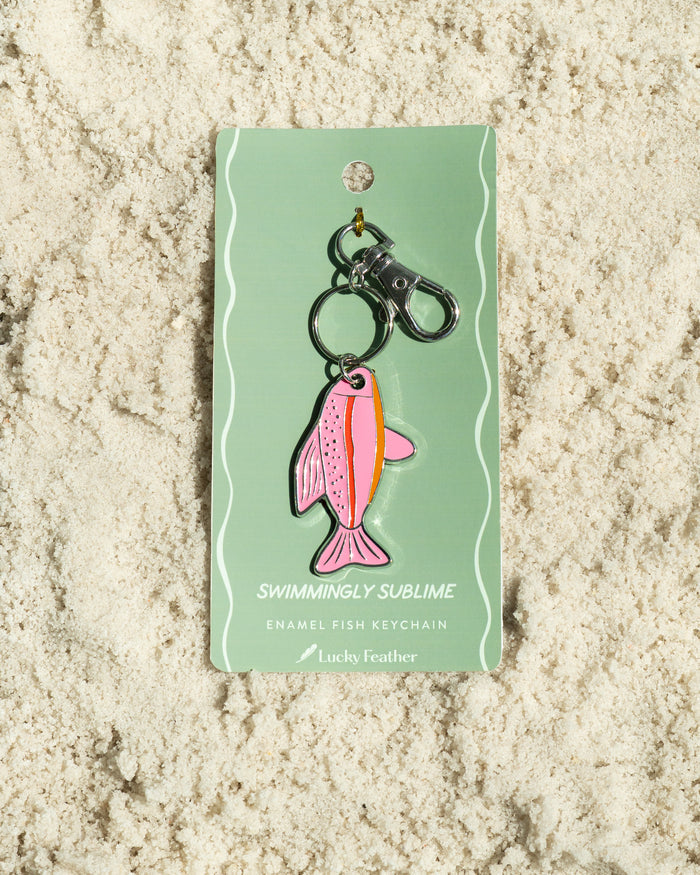 A pink enamel fish keychain from 'Lucky Feather' is displayed on a light green card on a bed of light-colored sand.