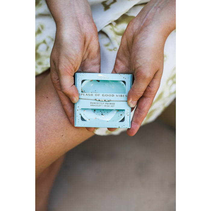 A person holds a blue gift box labeled 'Splash of Good Vibes Perfectly Paired Bracelet + Dish Set'.