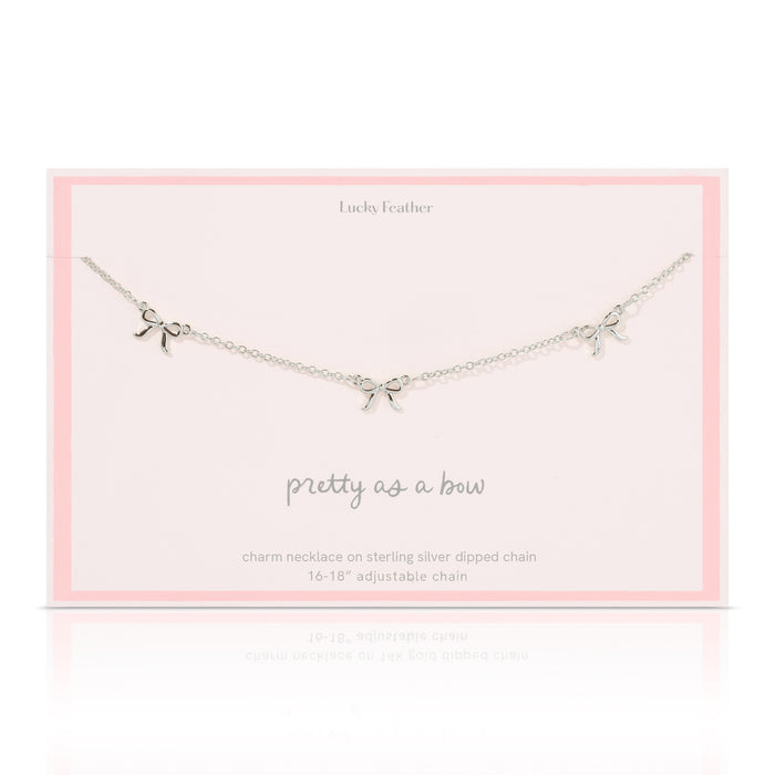 A silver-colored necklace with three bow charms lies on a pale pink card from 'Lucky Feather'.