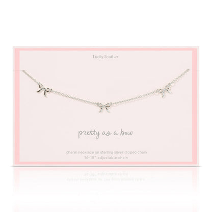 A silver-colored necklace with three bow charms lies on a pale pink card from 'Lucky Feather'.