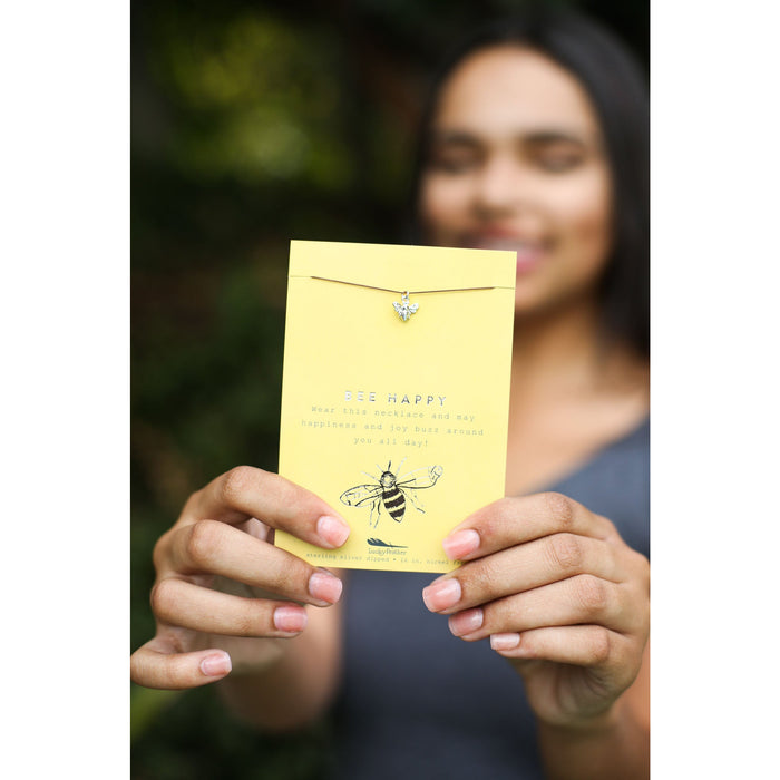 A woman holds a 'Bee Happy' necklace on a yellow card.