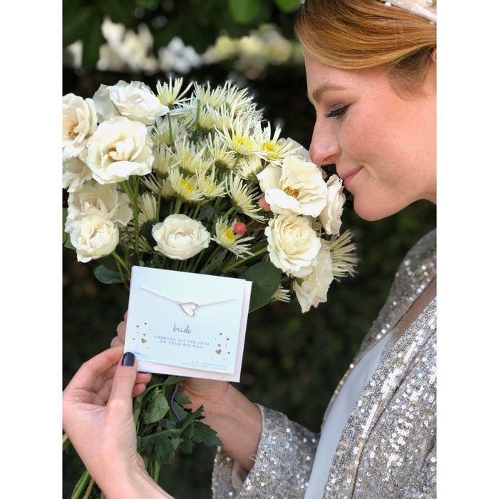 A woman smelling a bouquet of white roses and daisies holds a card featuring a necklace that reads 'bride.'
