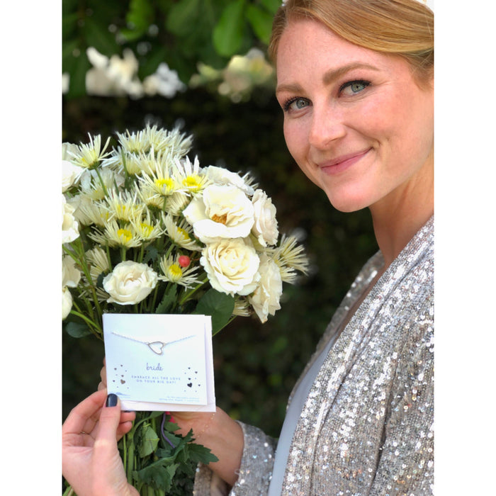 A smiling woman in a sequined jacket holds a bouquet of flowers and a 'Bride' necklace card.