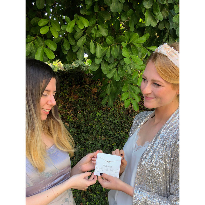 Two women stand outdoors in front of greenery, one handing the other a card that reads 'bridesmaid.'