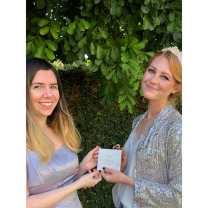 Two women pose in front of foliage, holding a gift card that reads 'bridesmaid.'