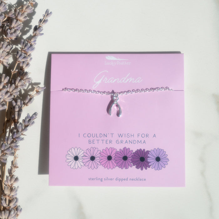 A silver necklace with a wishbone charm rests on a pink card with the text 'Grandma' and an arrangement of daisies, next to dried lavender.