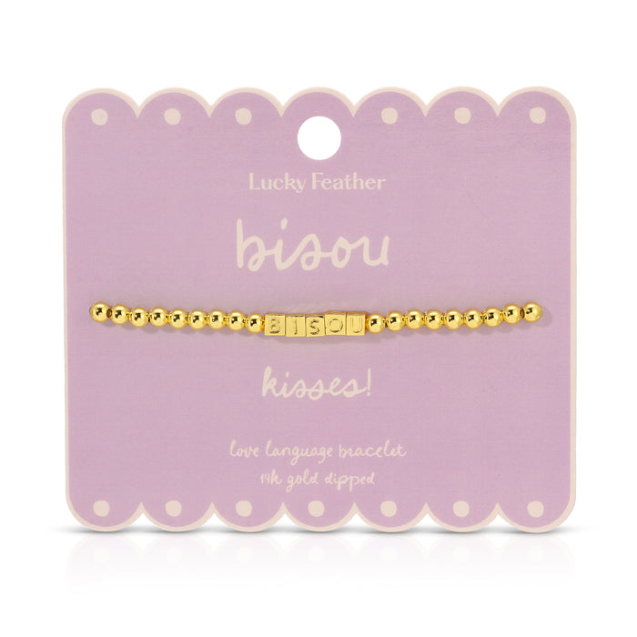 A gold beaded bracelet with the word 'bisou' spelled out in square letter beads is displayed on a lavender card.