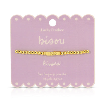 A gold beaded bracelet with the word 'bisou' spelled out in square letter beads is displayed on a lavender card.