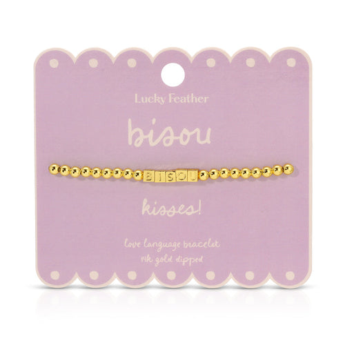 A gold beaded bracelet with the word 'bisou' spelled out in square letter beads is displayed on a lavender card.