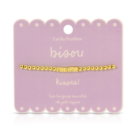 A gold beaded bracelet with the word 'bisou' spelled out in square letter beads is displayed on a lavender card.