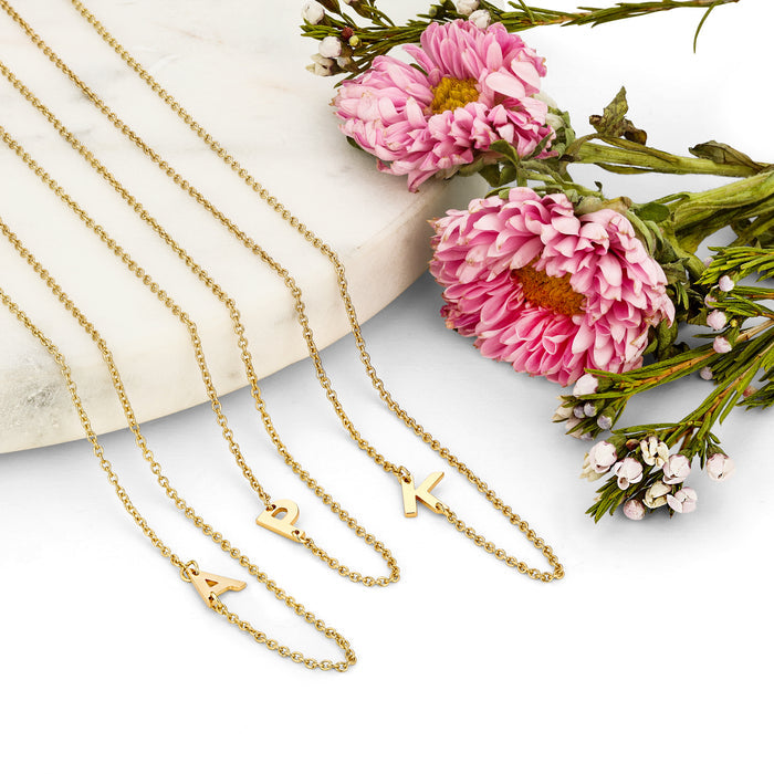 Three gold initial necklaces are displayed with pink and white flowers on a marble-like surface.