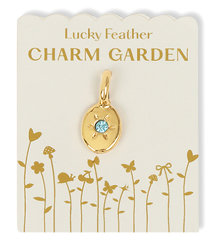 A gold-colored charm with a blue gem sits on a 'Lucky Feather Charm Garden' display card with drawings of flowers, insects, and other flora.