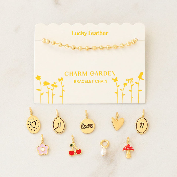 A gold ball chain bracelet displayed on a card labeled 'Lucky Feather Charm Garden Bracelet Chain' with assorted gold charms scattered below.