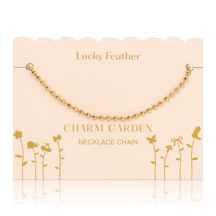 A gold-colored ball chain necklace is displayed on a peach-colored card with the words 'Lucky Feather Charm Garden Necklace Chain' and gold floral silhouette graphics.