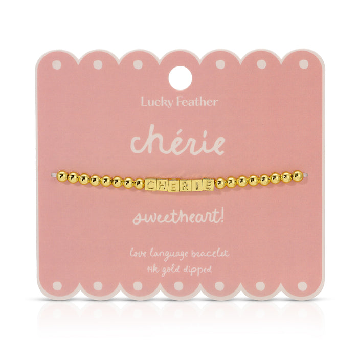 A 'ch√©rie' bracelet with gold beads and lettered blocks is displayed on a scalloped pink card by Lucky Feather.