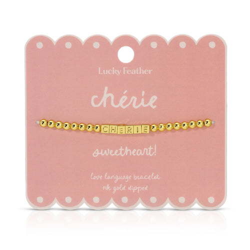 A 'ch√©rie' bracelet with gold beads and lettered blocks is displayed on a scalloped pink card by Lucky Feather.