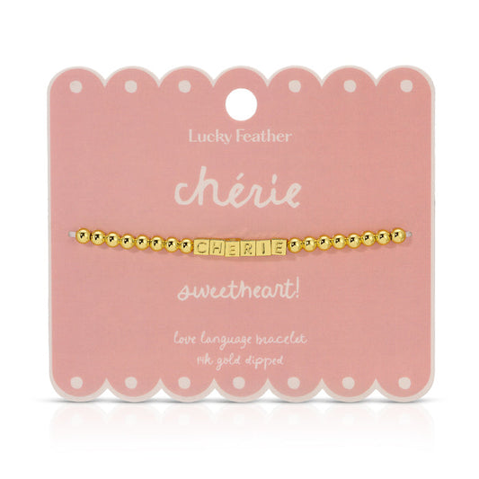 A 'ch√©rie' bracelet with gold beads and lettered blocks is displayed on a scalloped pink card by Lucky Feather.