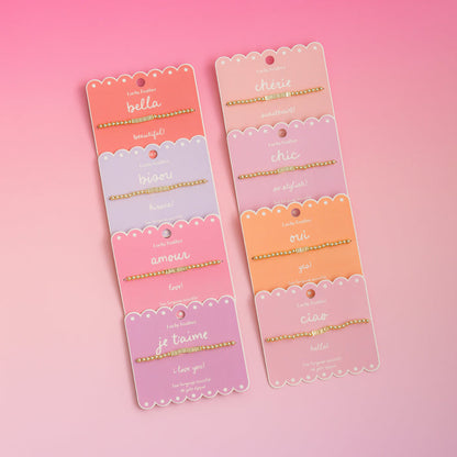 Several 'Lucky Feather' beaded bracelets with different words are displayed on pink scalloped cards against a pink background.