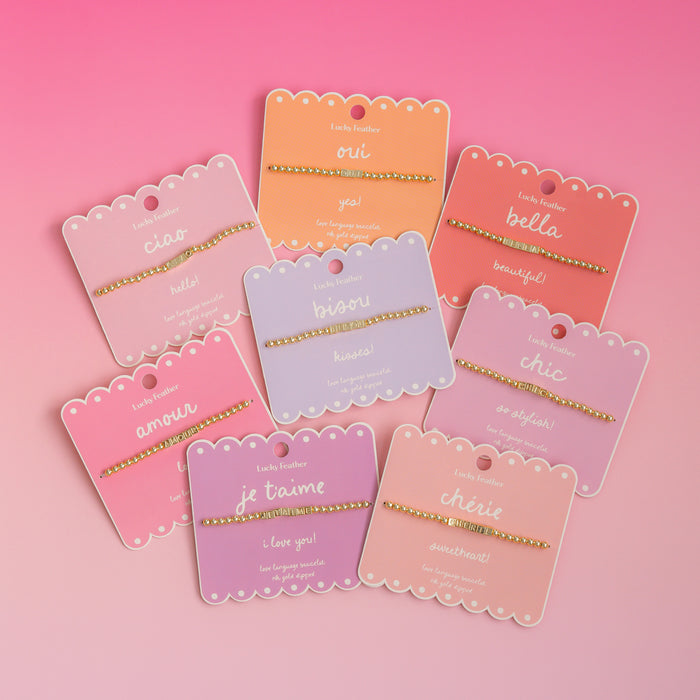 Several 'Lucky Feather' bracelets with different words in various pastel-colored packaging are displayed on a pink background.