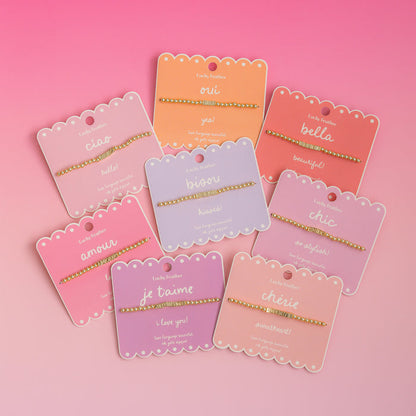 Several 'Lucky Feather' bracelets with different words in various pastel-colored packaging are displayed on a pink background.