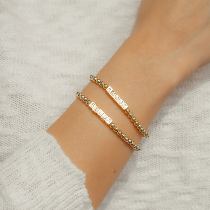 A person's arm wearing two beaded gold bracelets with the words 'BELLA' and 'AMOUR'.
