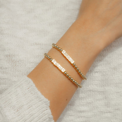 A person's arm wearing two beaded gold bracelets with the words 'BELLA' and 'AMOUR'.