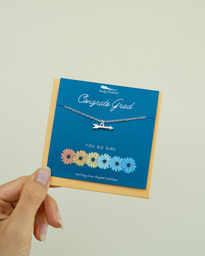A hand holds a 'Congrats Grad' card with a silver arrow necklace and daisy illustrations.