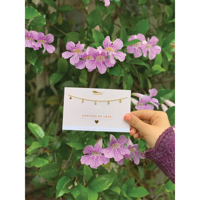 A hand holds a 'Bunches of Love' necklace card against a backdrop of purple flowers and green leaves.