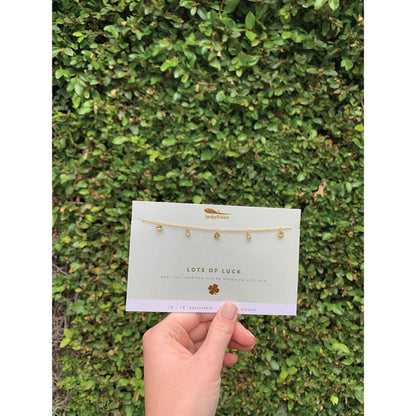 A hand holds a 'Lots of Luck' necklace by Imoky Feather against a green bush background.