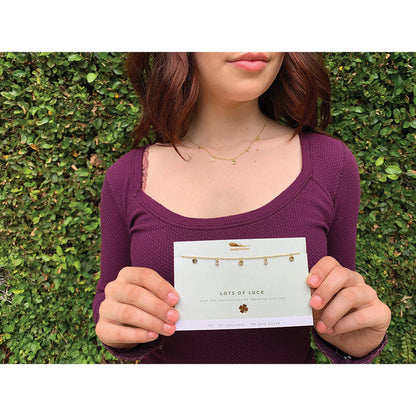 A woman wears a delicate gold necklace and holds a card displaying the same necklace labeled 'Lots of Luck.'