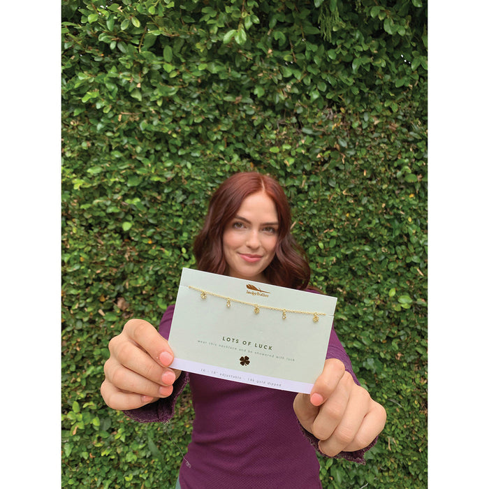 A woman holds up a 'Lots of Luck' necklace package against a backdrop of greenery.