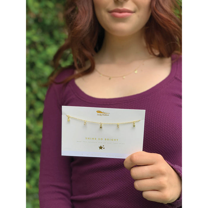 A woman holds a card displaying a 'Shine So Bright' necklace by Irmaky Feather, featuring star and jewel pendants.