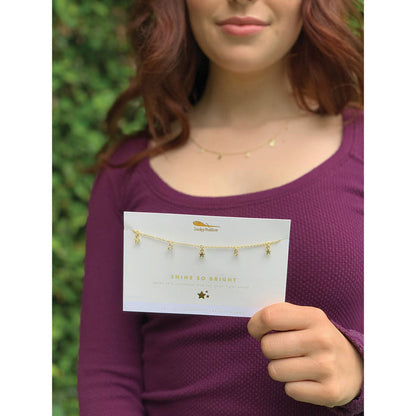 A woman holds a card displaying a 'Shine So Bright' necklace by Irmaky Feather, featuring star and jewel pendants.