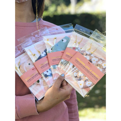 A person holds several individually packaged 'Lucky Feather Spreading Love & Kindness' face masks with gold chain holders.
