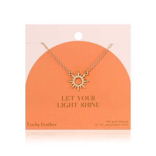 A gold-colored sun pendant hangs on a chain from a peach-colored display card that reads 'Let Your Light Shine'.