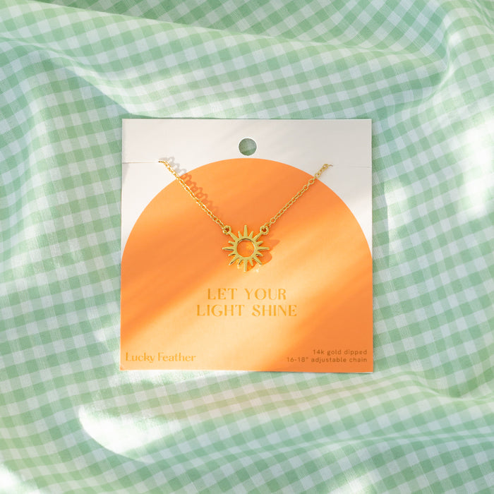 A gold-colored sun pendant necklace on a card that reads 'Let Your Light Shine,' displayed on a light green and white checkered fabric.