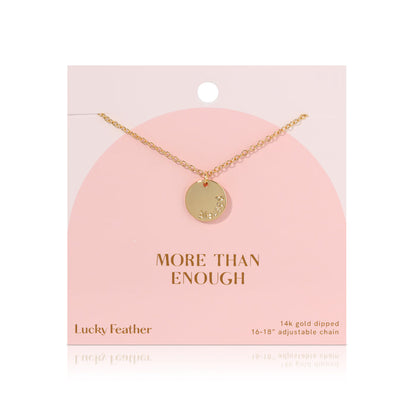 A 14k gold dipped necklace with a round pendant hangs against a pink card with 'MORE THAN ENOUGH' printed on it.