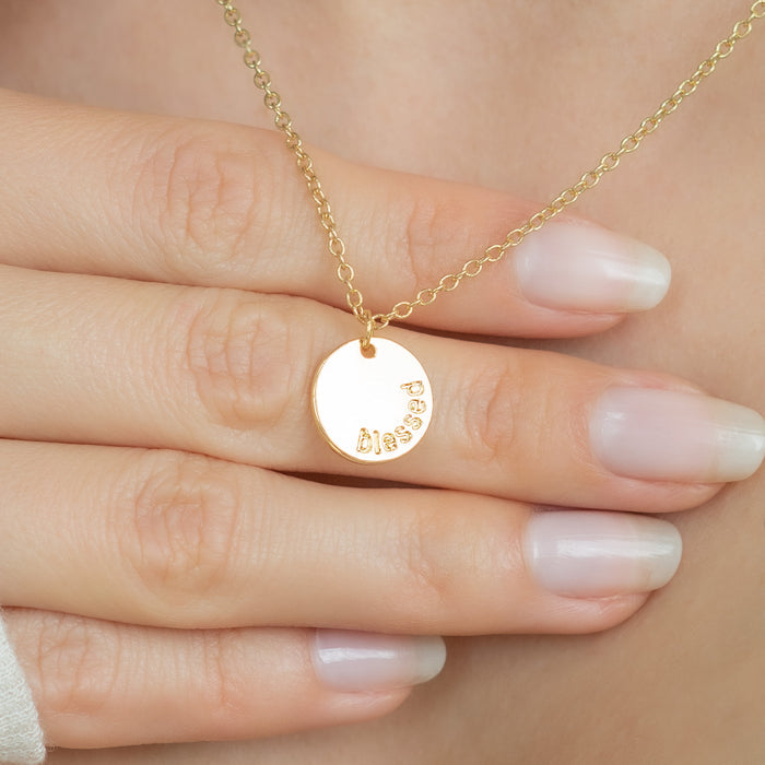 A hand is displaying a gold necklace with a circular pendant engraved with the word 'blessed.'