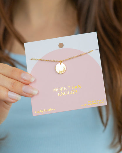 A hand holds a 'Lucky Feather' necklace and its pink display card that reads, 'More than Enough.'