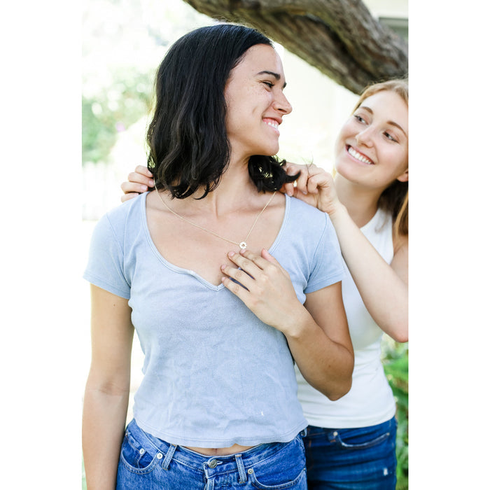 A person with dark hair adjusts a necklace on a smiling person wearing a blue shirt.