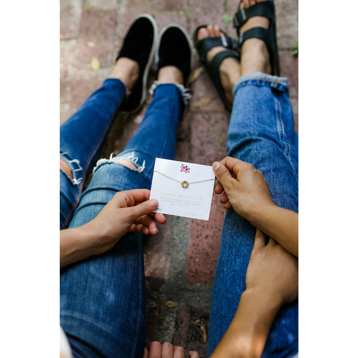 Two people wearing jeans and shoes hold a 'Flowers Will Bloom' necklace.