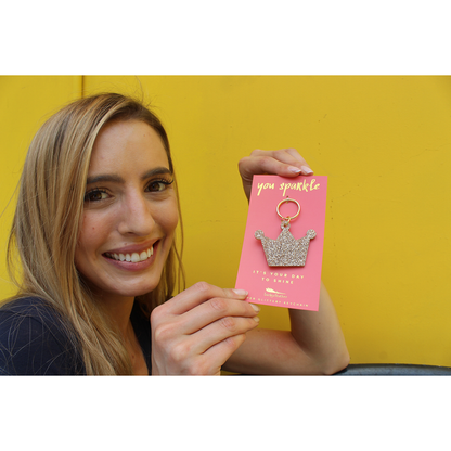 A woman smiles while holding up a 'you sparkle' card with a glittery crown keychain against a yellow background.