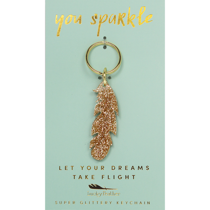 A gold glitter feather keychain hangs on a light blue display card.