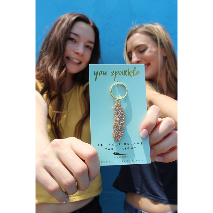 Two smiling women hold up a 'Lucky Feather' brand sparkly feather keychain against a blue background.
