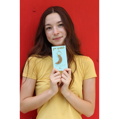 A young woman in a yellow shirt holds up a 'You Sparkle' card with a crescent moon keychain.